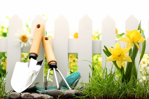 A close-up view of a small garden scene showing two gardening tools—a trowel and a hand fork—resting on a patch of dark soil next to bright yellow daffodils and white daisies with yellow centers, set against a white picket fence background. The garden features a lush, green grass lawn, with the tools positioned in the foreground, indicating recent or ongoing gardening activity. The scene is illuminated by natural sunlight, creating a cheerful and inviting outdoor environment typical of a well-maintained private garden in Bayswater or surrounding areas. The overall composition emphasizes garden maintenance elements and seasonal flower growth, aligning with professional gardening and hedge trimming services offered by Hedge Trimming Bayswater, providing visual context for outdoor lawn and planting care in the local area.